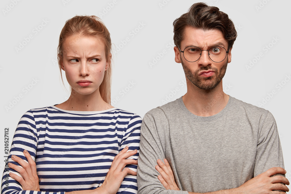 Indoor shot of grumpy European couple have displeased expressions after quarrel, don`t speak to each other, look angrily, keep hands crossed can`t find common solution, isolated on white wall Indoor shot of grumpy European couple have displeased expressions after quarrel, don`t speak to each other, look angrily, keep hands crossed can`t find common solution, isolated on white wall