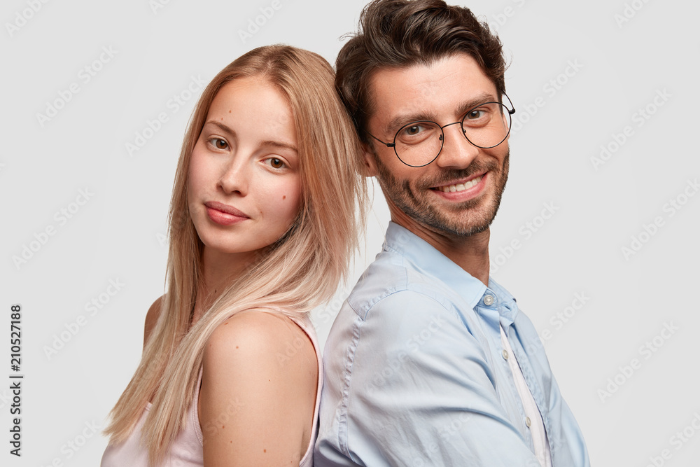 Couple stands backs, look positively at camera, enjoy togetherness and closeness, feel support from each other, pose against white concrete wall. Cute young European woman stands near boyfriend Couple stands backs, look positively at camera, enjoy togetherness and closeness, feel support from each other, pose against white concrete wall. Cute young European woman stands near boyfriend