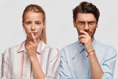 Serious cute Caucasian female and her young male partner have thoughtful expressions, try to concentrate, find solution, dressed in shirts, isolated over white background. Partnership concept