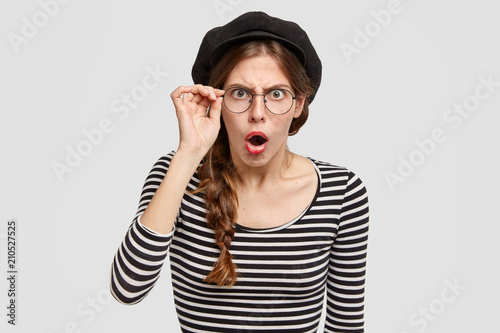 Photos Female from France in stupor, keeps mouth opened, has angry surprised expression as notices some default on work, dressed in fashionable retro outfit