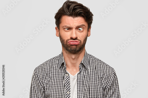 Funny European male makes grimace, purses lips and crosses eyes, foolishes indoor, has fun, dressed in fashionable shirt, isolated over white background. People and facial expressions concept photo