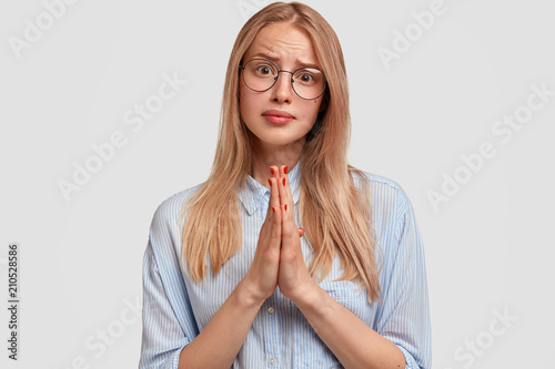 Adorable lovely female asks for apology, has pleading expression, keeps palms pressed together, wears spectacles, hopes to get permission from mother to go on party with friends. Begging concept