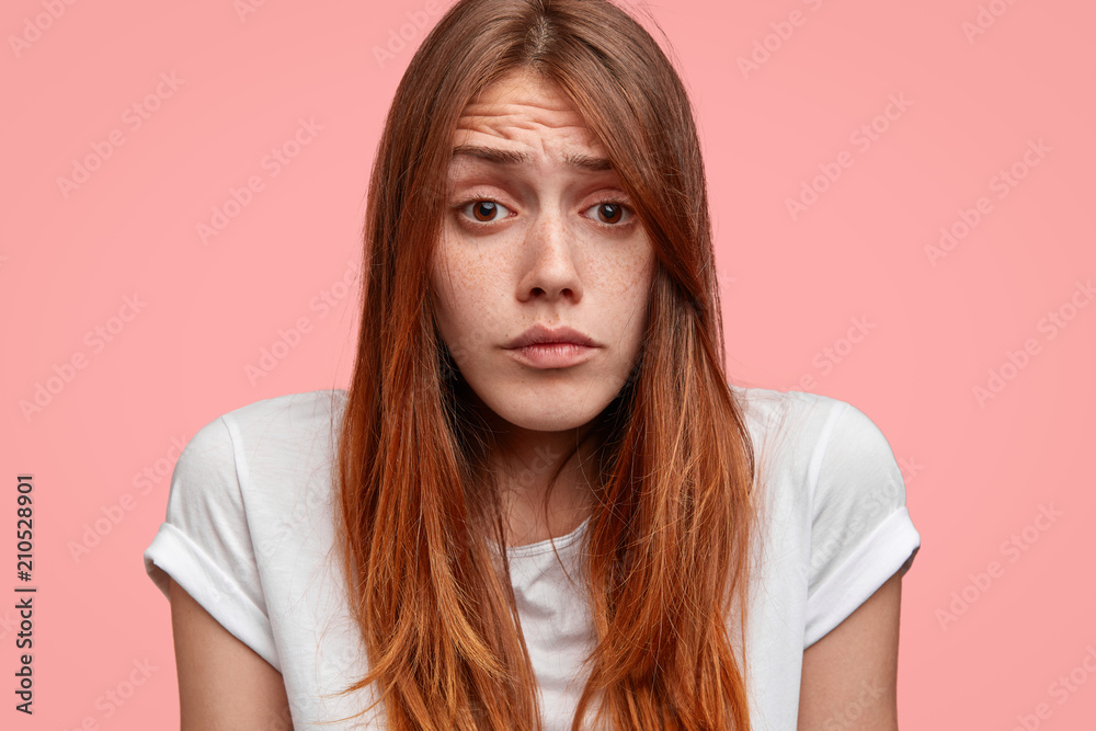 Close up portrait of attractive freckled female with sorrowful expression, looks pity, need support and help in difficult situation, dressed in casual white t shirt, stands against pink background