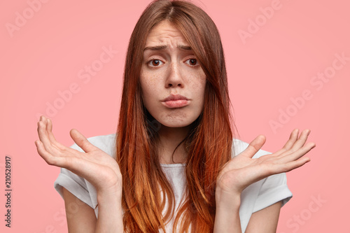 Confused adorable freckled female has hesitant expression, shrugs shoulders, curves lips, has long hair, dressed in casual clothing, stands against pink background. Puzzled European woman uncertain