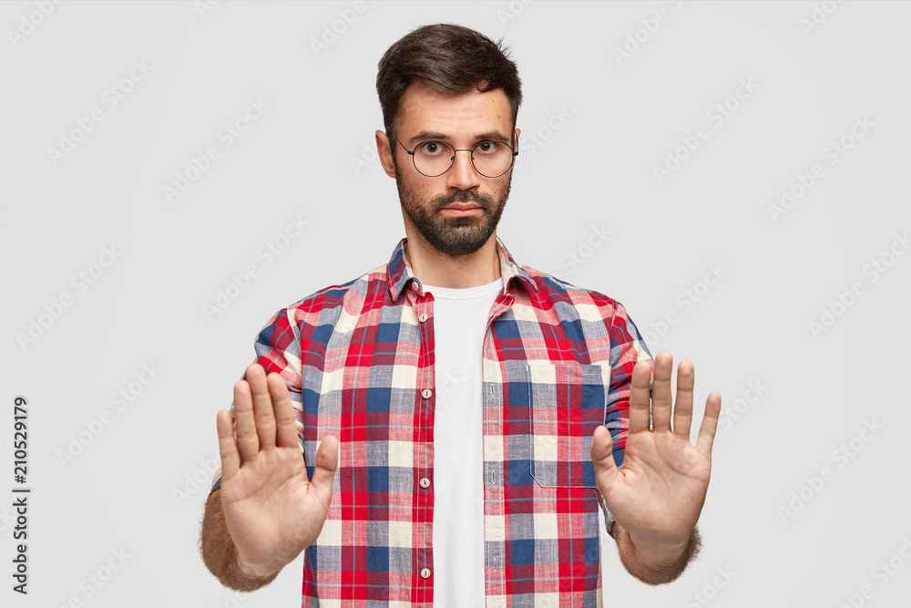 Fototapeta premium Pleasant looking serious male office worker refuses to help with preparing project, shows stop gesture, asks not bother him during vacation, has thick beard and mustache, isolated on white wall
