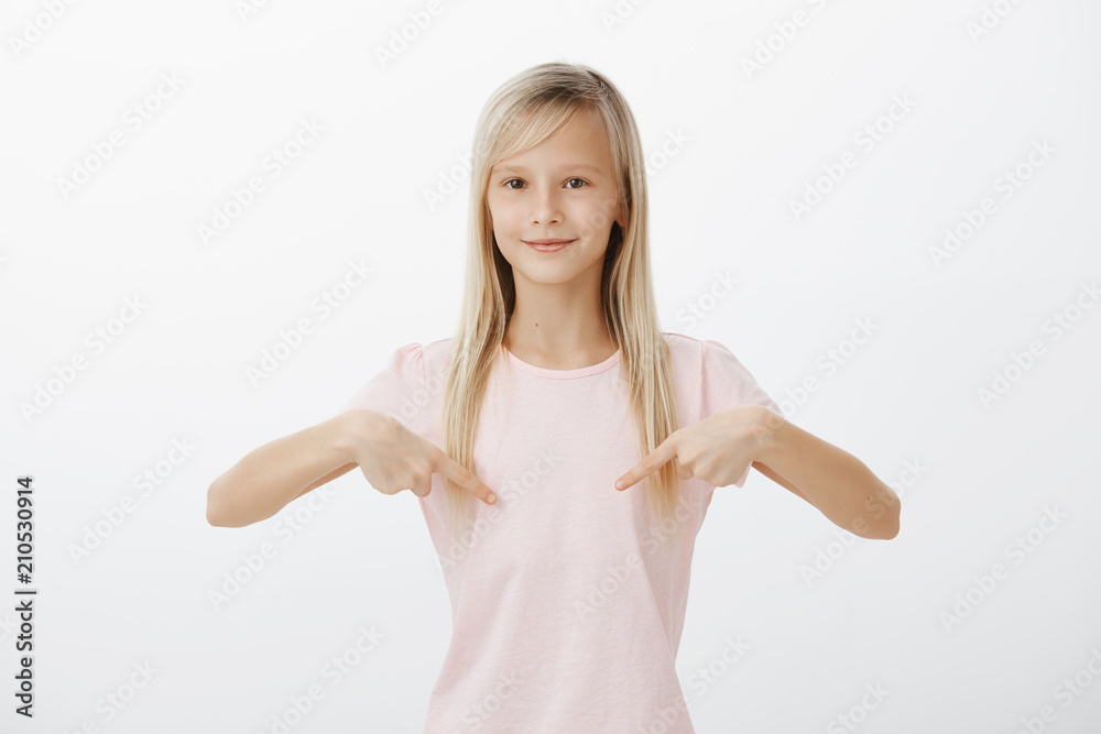 Portrait of proud fashionable girl with blond hair pointing at pink t-shirt with pleased and happy expression, showing girls in class new outfit, standing against gray background Portrait of proud fashionable girl with blond hair pointing at pink t-shirt with pleased and happy expression, showing girls in class new outfit, standing against gray background