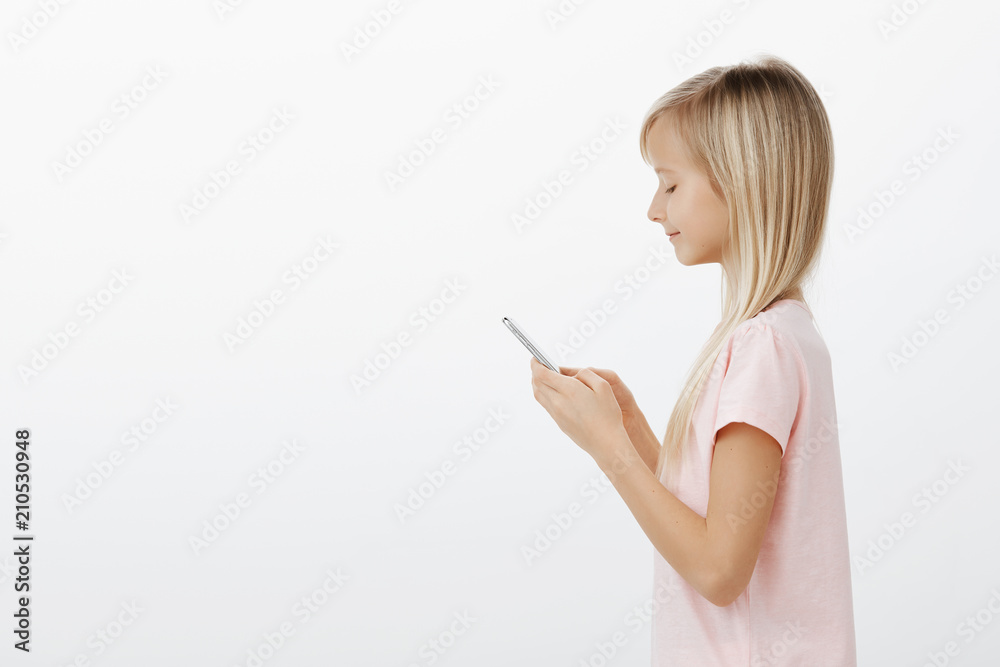 Profile portrait of joyful calm attractive girl with blond hair in pink t-shirt, holding smartphone, smiling at screen, sending photo to mom via messages, telling mom she finished house duties Profile portrait of joyful calm attractive girl with blond hair in pink t-shirt, holding smartphone, smiling at screen, sending photo to mom via messages, telling mom she finished house duties