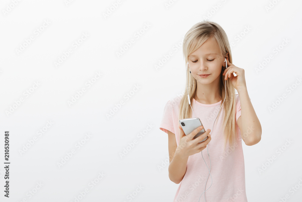 Smart girl knows everything about gadgets. Portrait of cute beautiful blond young child, wearing earphones and picking song in smartphone, looking at screen focused, standing over gray background Smart girl knows everything about gadgets. Portrait of cute beautiful blond young child, wearing earphones and picking song in smartphone, looking at screen focused, standing over gray background