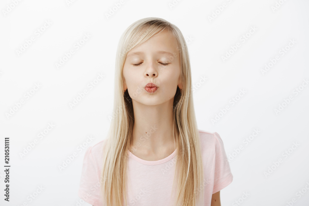 Girl practising near mirror how to kiss. Portrait of cute fashionable young girl with blond hair, folding lips and closing eyes while waiting for mwah, dreaming and standing over gray background Girl practising near mirror how to kiss. Portrait of cute fashionable young girl with blond hair, folding lips and closing eyes while waiting for mwah, dreaming and standing over gray background