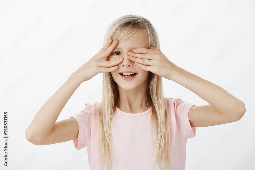Curious daughter wants to see what father prepared for b-day. Portrait of intrigued impatient cute girl with beautiful blond hair, covering eyes with palms and peeking with broad smile over gray wall Curious daughter wants to see what father prepared for b-day. Portrait of intrigued impatient cute girl with beautiful blond hair, covering eyes with palms and peeking with broad smile over gray wall