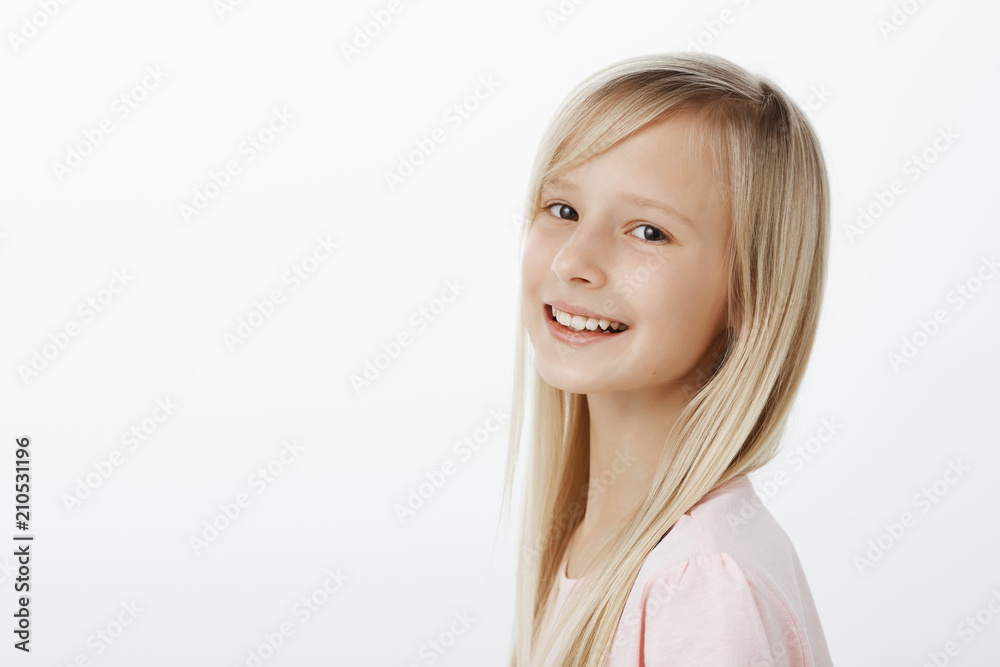 Profile shot of satisfied happy little caucasian girl with long blond hair, turning at camera and smiling cheerfully, having fun while walking with parents in park, spending great time over gray wall Profile shot of satisfied happy little caucasian girl with long blond hair, turning at camera and smiling cheerfully, having fun while walking with parents in park, spending great time over gray wall