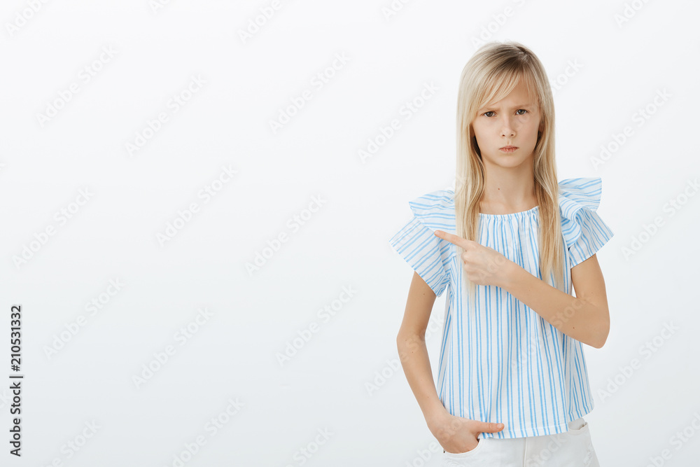 Deal with that boy. Portrait of offended angry european girl with blond hair, frowning and staring from under forehead, pointing at upper left corner while standing over gray wall upset Deal with that boy. Portrait of offended angry european girl with blond hair, frowning and staring from under forehead, pointing at upper left corner while standing over gray wall upset