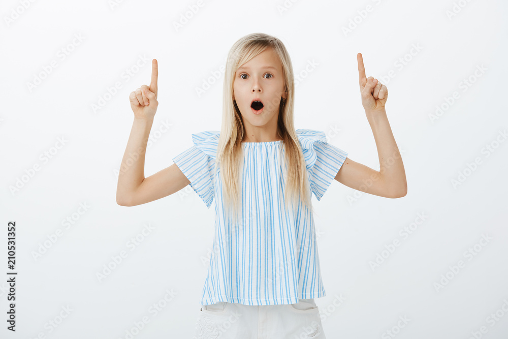 Portrait of surprised amazed cute young girl with fair hair, dropping jaw from wonder and pointing upwards, sharing stunning news with sister, being excited and thrilled over gray background Portrait of surprised amazed cute young girl with fair hair, dropping jaw from wonder and pointing upwards, sharing stunning news with sister, being excited and thrilled over gray background