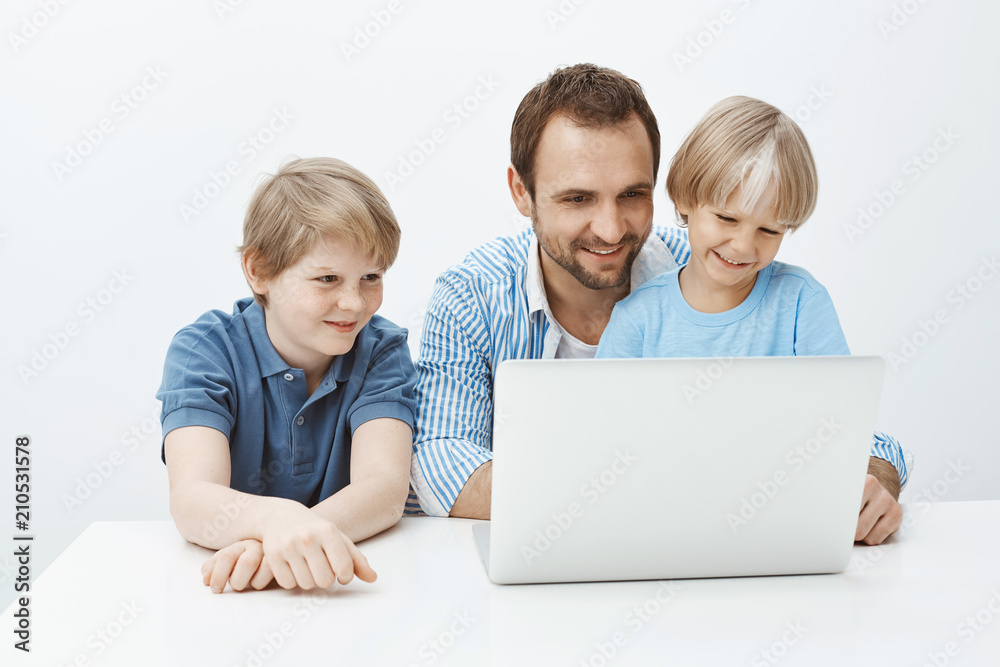 Dad recalling happy memories from summer vacation with sons. Portrait of satisfied carefree family of boys and father sitting at table near laptop, smiling broadly at screen over gray background Dad recalling happy memories from summer vacation with sons. Portrait of satisfied carefree family of boys and father sitting at table near laptop, smiling broadly at screen over gray background
