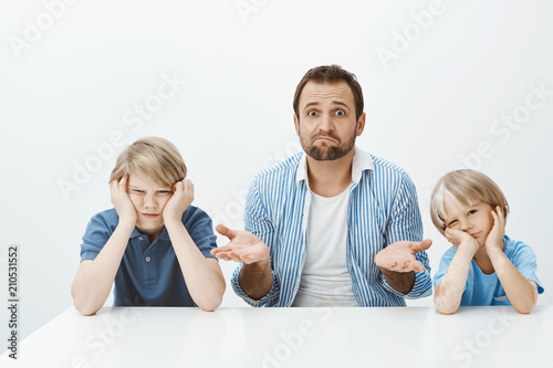 Why sons are mad. Portrait of clueless nervous father-in-law sitting with two cute boys at table, shrugging and being questioned how lift mood while guys are bored and indifferent over gray wall