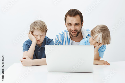 Pleased funny father sitting with sons near laptop, watching at screen with satisfied happy smile while boys feeling bored and indifferent, wanting play instead of spending time with dad