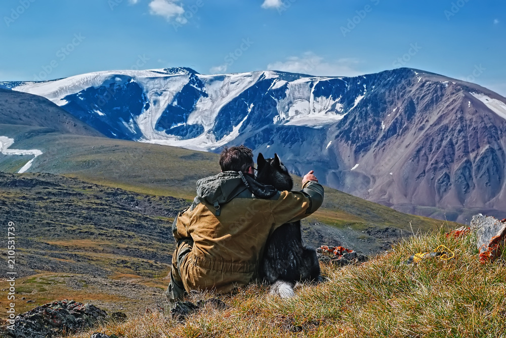 Concept of traveling with dog in the mountains. Man with Siberian husky looks at the snow-capped mountains. Back view.