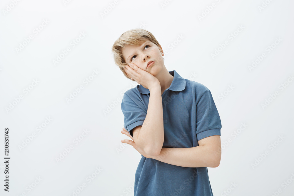 Indoor shot of displeased bored cute blond boy, leaning head on palm and staring up, frowning and feeling gloomy while being punished for disobedience, feeling boredom and negative emotions Indoor shot of displeased bored cute blond boy, leaning head on palm and staring up, frowning and feeling gloomy while being punished for disobedience, feeling boredom and negative emotions