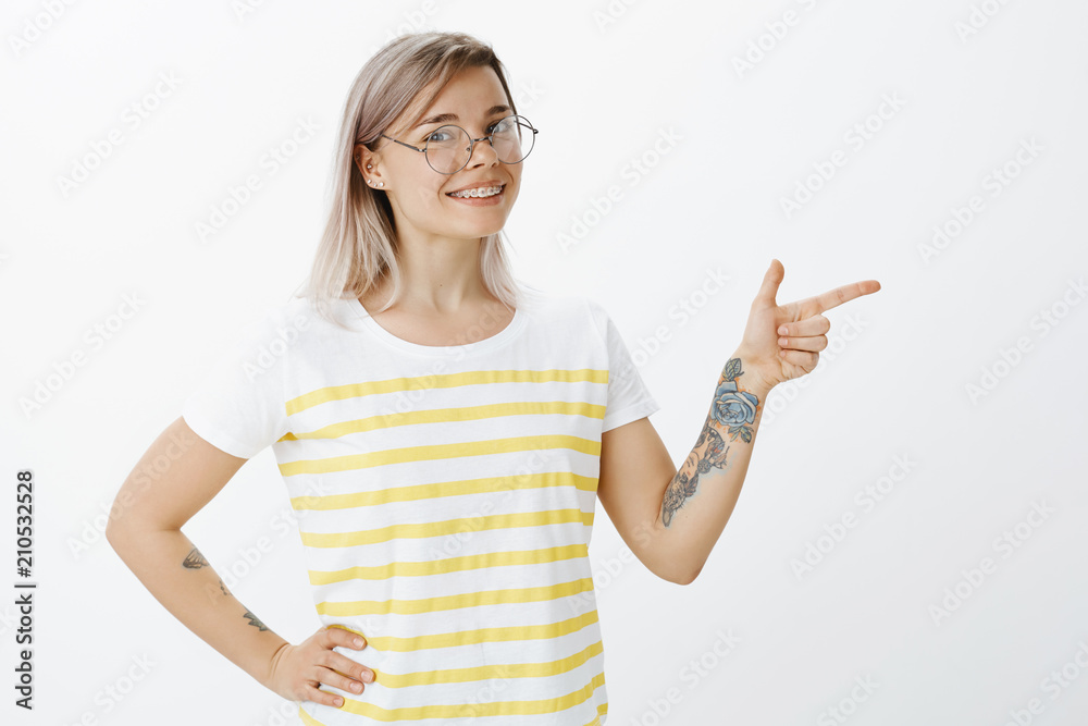 Check out, you shoud see this. Charming positive european female with fair hair and tattoos, pointing right with finger gun gesture, holding hand on hip and smiling broadly while giving direction Check out, you shoud see this. Charming positive european female with fair hair and tattoos, pointing right with finger gun gesture, holding hand on hip and smiling broadly while giving direction