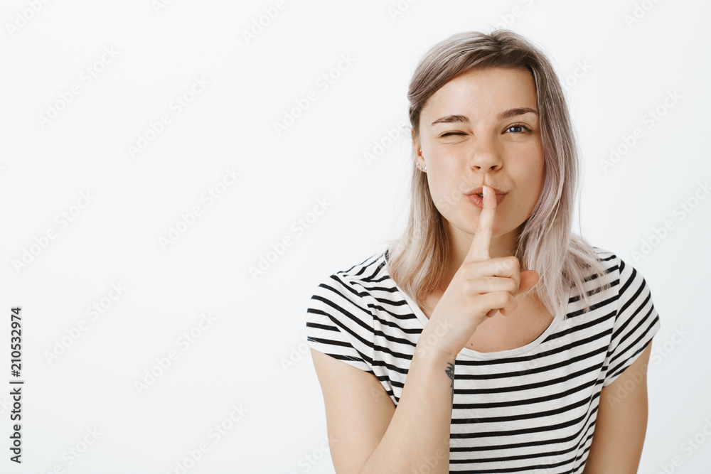 Do not tell anyone what happened. Portrait of attractive playful stylish girl, bending towards camera, winking as if hinting on secret and saying shh while showing shush gesture, gossiping or rumoring Do not tell anyone what happened. Portrait of attractive playful stylish girl, bending towards camera, winking as if hinting on secret and saying shh while showing shush gesture, gossiping or rumoring