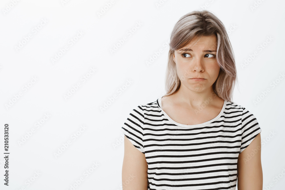 Waist-up shot of lonely gloomy cute girlfriend with fair hair in striped t-shirt, sulking and frowning, looking left, being upset and offended, expressing sadness while standing against gray wall Waist-up shot of lonely gloomy cute girlfriend with fair hair in striped t-shirt, sulking and frowning, looking left, being upset and offended, expressing sadness while standing against gray wall