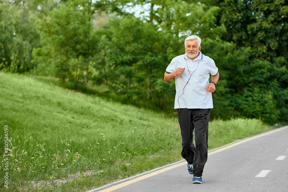 Fit old man running on racetrack in green park. Outdoors sport, trainings.  Wearing classic white polo shirt with dark blue stripes,black trousers,  sneakers. Listening to music with red headphones. Stock Photo