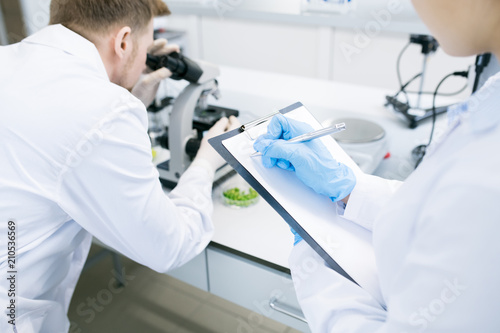 Crop back view of female microbiologist writing down into clipboard data received from man analyzing green vegetable structure under microscope