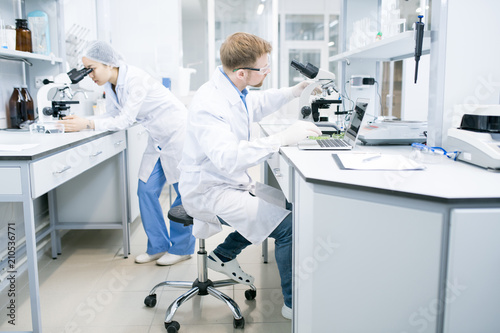 Side view of male microbiologist in glasses sitting at desk using laptop and holding hand on microscope with food samples with assistant looking at microscope eyepiece on background .