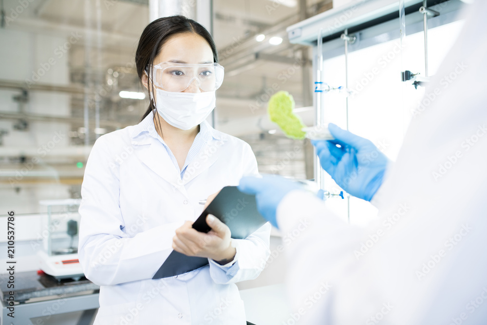 Crop back view of microbiologist holding in hands pincers with green food nutrition test sample and woman looking at it writing down results into clipboard Crop back view of microbiologist holding in hands pincers with green food nutrition test sample and woman looking at it writing down results into clipboard