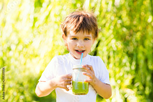 Cute little boy drinks healthy green smoothie with straw in a jar mug against the background of greenery outdoor. .