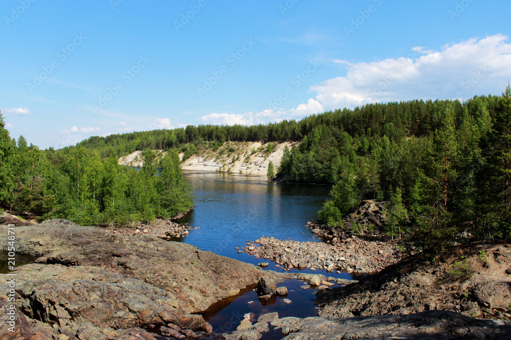 Fototapeta premium A wide river flows between sandy beaches surrounded by a mixed coniferous forest. Karelia, Russia. In the foreground is an extinct volcano called Girvas with black stones