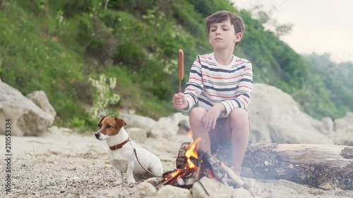 Slow motion a happy boy is sitting on a log with his dog breed Jack Russell and frying on a campfire marshmallow on the seashore against the backdrop of the setting sun. A child with his pet