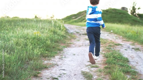 A happy boy runs along a country road through a beautiful landscape of green hills at sunset on a sunny day. Back view