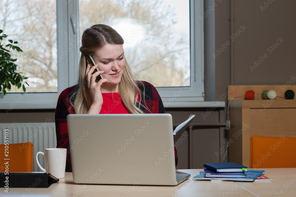 A blonde woman sitting at a laptop and talking over a cell phone. A blonde woman sitting at a laptop and talking over a cell phone.