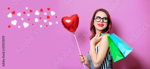 Young red-haired woman with heart shape toy and shopping bags on yellow background