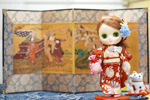 Cute doll in Kimono. A cute doll is dressing up in a red kimono. Kimono is a traditional Japanese garment. The doll is standing in Japanese setting holding omamori Japanese charm.