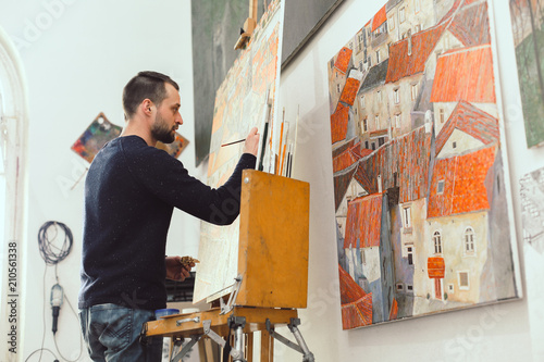 Concentrated male artist is drawing on easel in his art studio, lots of pictures, creative atmosphere, side view, space for text