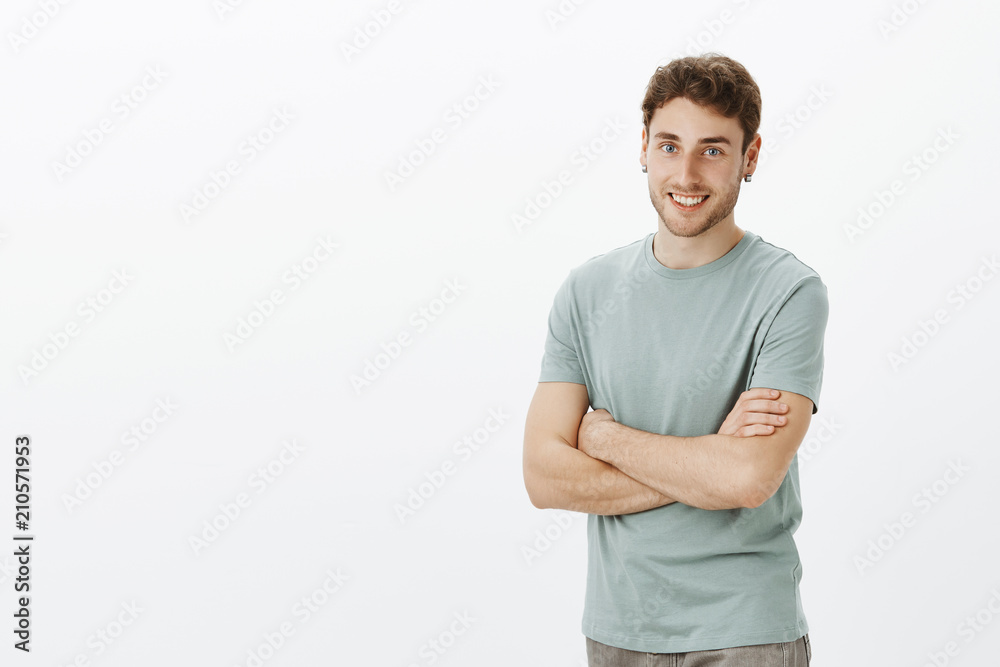 Portrait of friendly excited handsome man with fair hair and earrings, standing half-turned with crossed arms over chest and smiling while listening to daily task from boss over gray wall Portrait of friendly excited handsome man with fair hair and earrings, standing half-turned with crossed arms over chest and smiling while listening to daily task from boss over gray wall