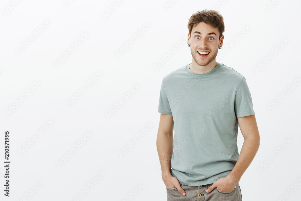 Guy hearing amazing news, feeling happy for friend. Portrait of positive friendly-looking handsome man in t-shirt, holding hands in pockets and smiling broadly hearing surprise or awesome story Guy hearing amazing news, feeling happy for friend. Portrait of positive friendly-looking handsome man in t-shirt, holding hands in pockets and smiling broadly hearing surprise or awesome story