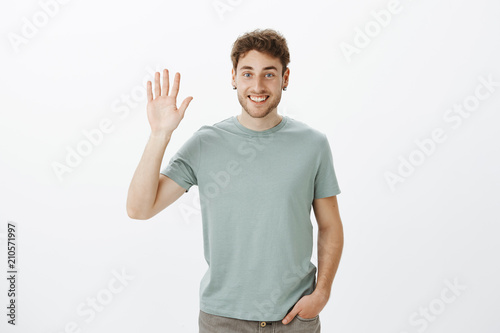 Hi nice to meet you. Portrait of handsome outgoing european guy in casual t-shirt raising hand and waving palm in hello gesture, greeting friend and smiling broadly while standing over gray wall