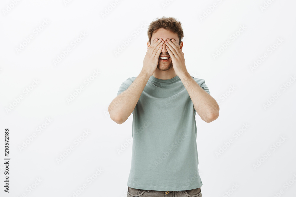 Indoor shot of playful good-looking mature man playing hide and seek with children smiling broadly while covering eyes and face with palm, waiting over gray background and counting Indoor shot of playful good-looking mature man playing hide and seek with children smiling broadly while covering eyes and face with palm, waiting over gray background and counting