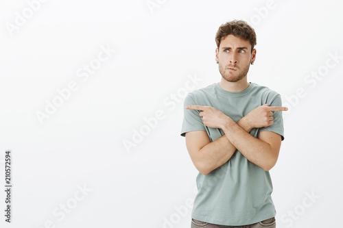 Portrait of confused european male model in earrings, smirking and frowning while thinking, crossing hands and pointing left and right while choosing direction, standing and deciding where to go