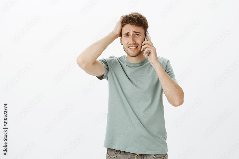 Troubled upset good-looking male model with fair hair, frowning and clenching teeth, touching hair, holding smartphone near ear, hearing terrible news and being sad, feeling miserable over grey wall Troubled upset good-looking male model with fair hair, frowning and clenching teeth, touching hair, holding smartphone near ear, hearing terrible news and being sad, feeling miserable over grey wall
