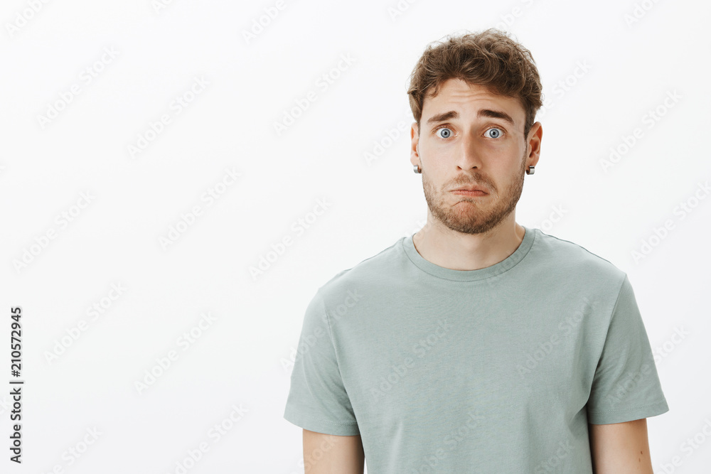 Indoor shot of intense sad and cute guy with beard and earrings, lifting eyebrow and pulling smile down, being miserable and upset with failed mission, standing gloomy over grey wall