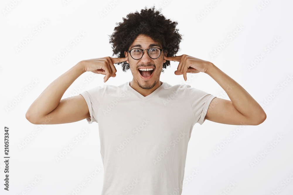 Carefree good-looking funny hispanic male model with curly hair, closing ears with index fingers and shouting, being in loud pub, watching football match, standing in noisy room over grey wall Carefree good-looking funny hispanic male model with curly hair, closing ears with index fingers and shouting, being in loud pub, watching football match, standing in noisy room over grey wall