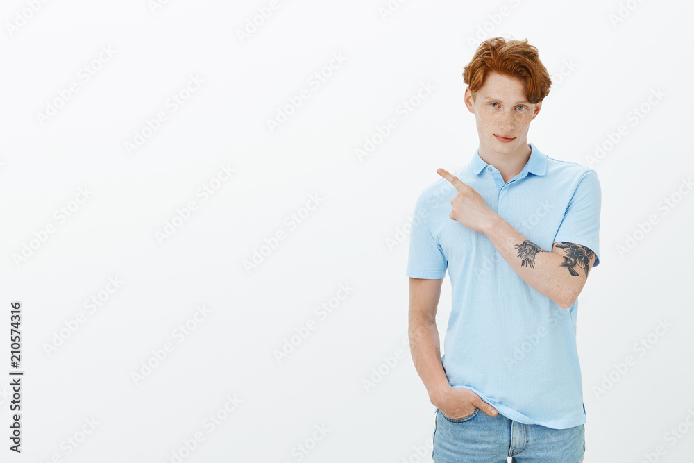 Confident handsome macho man with red hair, freckles and tatooed arm, pointing at upper left corner and gazing with slight smile at camera, being self-assured while giving direction over gray wall Confident handsome macho man with red hair, freckles and tatooed arm, pointing at upper left corner and gazing with slight smile at camera, being self-assured while giving direction over gray wall