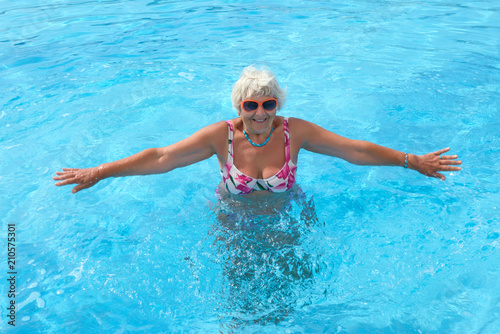 Aged woman is doing spa exercises in bright blue water of pool.
