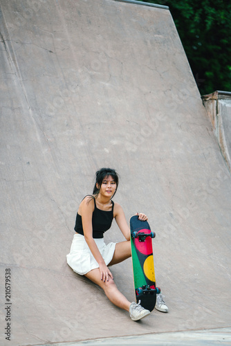 A charming Chinese Asian skater girl sits and lounges with her skateboard at a skate park on a sunny day. She enjoyed herself while riding on her skateboard the whole morning.