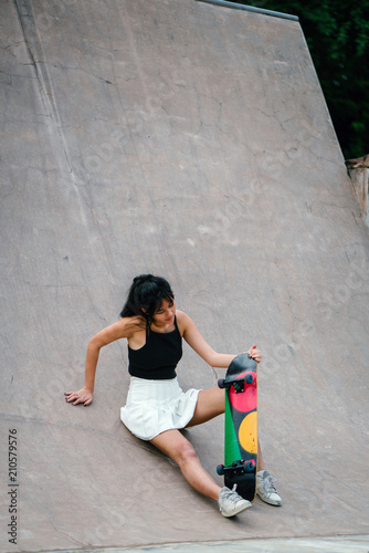 A playful Chinese Asian teenager sits with her skateboard at a skate park. She had a fun-filled afternoon riding her skateboard.