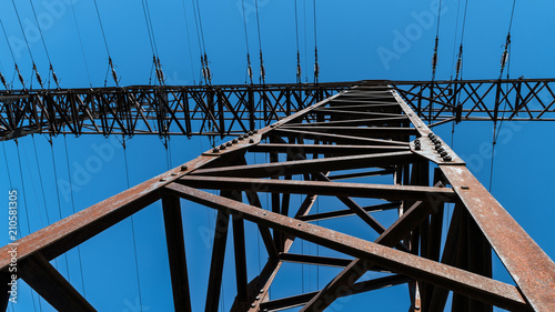 Transmission tower close-up against a blue sky. Silhouette of a tall rusty electricity pylon of power line from a frog's perspective. Idea of electric energy supply, transmission system, environment.